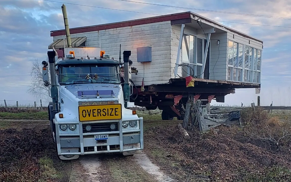 Mack truck carrying a weatherboard house along a rural gravel driveway